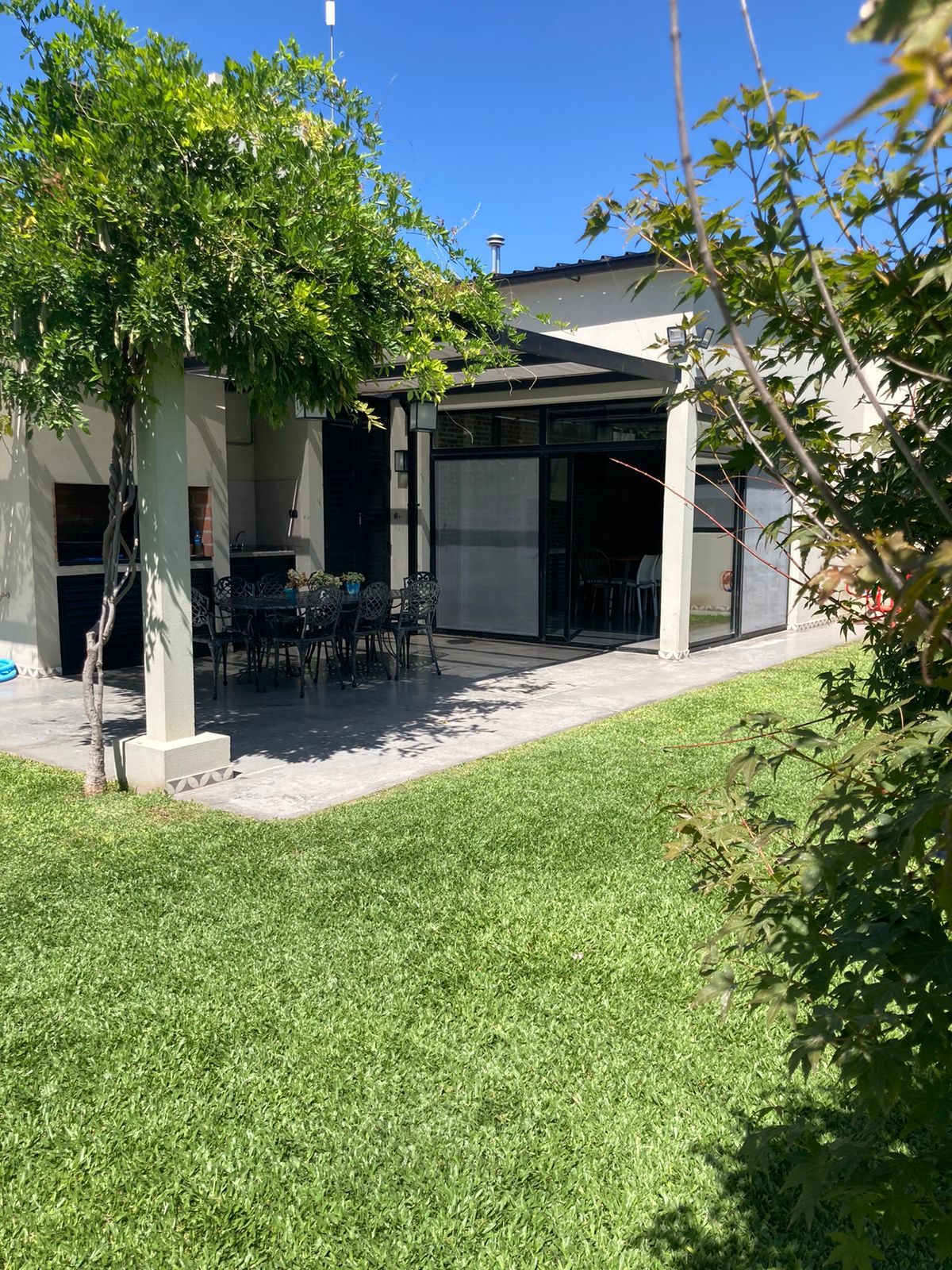 A spacious outdoor patio is visible, featuring a dining table surrounded by black decorative chairs. Lush green grass surrounds the area, complemented by trees providing gentle shade. The patio is enclosed by glass elements, inviting natural light into the space.