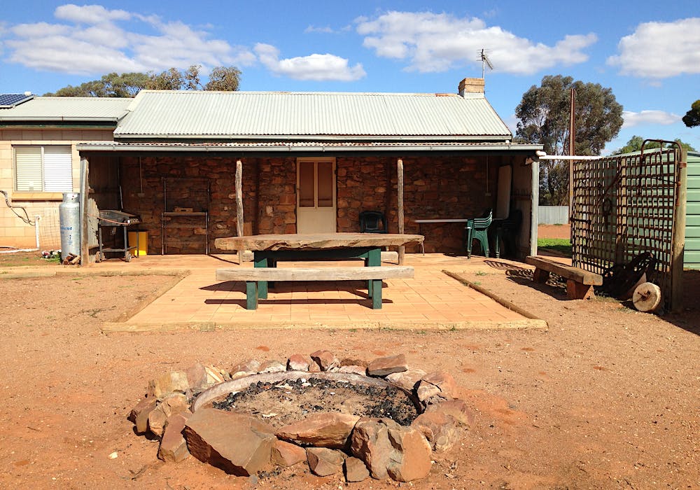 An outdoor area presents a spacious stone cottage with a corrugated metal roof. A large wooden table sits on a tiled patio, surrounded by red dirt. A simple fire pit is visible in the foreground, with surrounding stones arranged neatly. Lush greenery can be seen in the background.