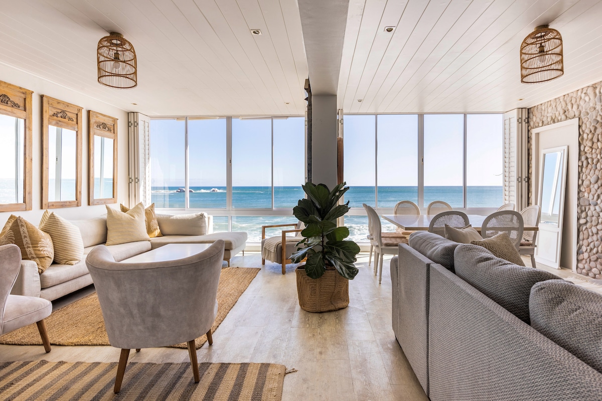 A spacious living area features large glass windows showcasing ocean views. Comfortable seating is arranged around a central plant. The decor includes natural elements like wooden accents and warm neutral tones, enhancing the beachside atmosphere, inviting relaxation and socializing.