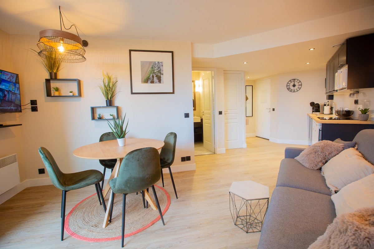 A modern dining area is arranged with a round wooden table surrounded by four green chairs. A cozy seating area is visible, featuring a gray sofa adorned with soft pillows. Wall-mounted plants and a decorative light fixture add visual interest to the space.