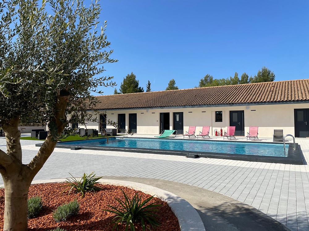 An outdoor area features a clear swimming pool surrounded by a spacious patio. Multiple lounge chairs in shades of pink are positioned alongside the water. Neatly trimmed landscaping includes an olive tree and decorative plants, with a building providing room access in the background.