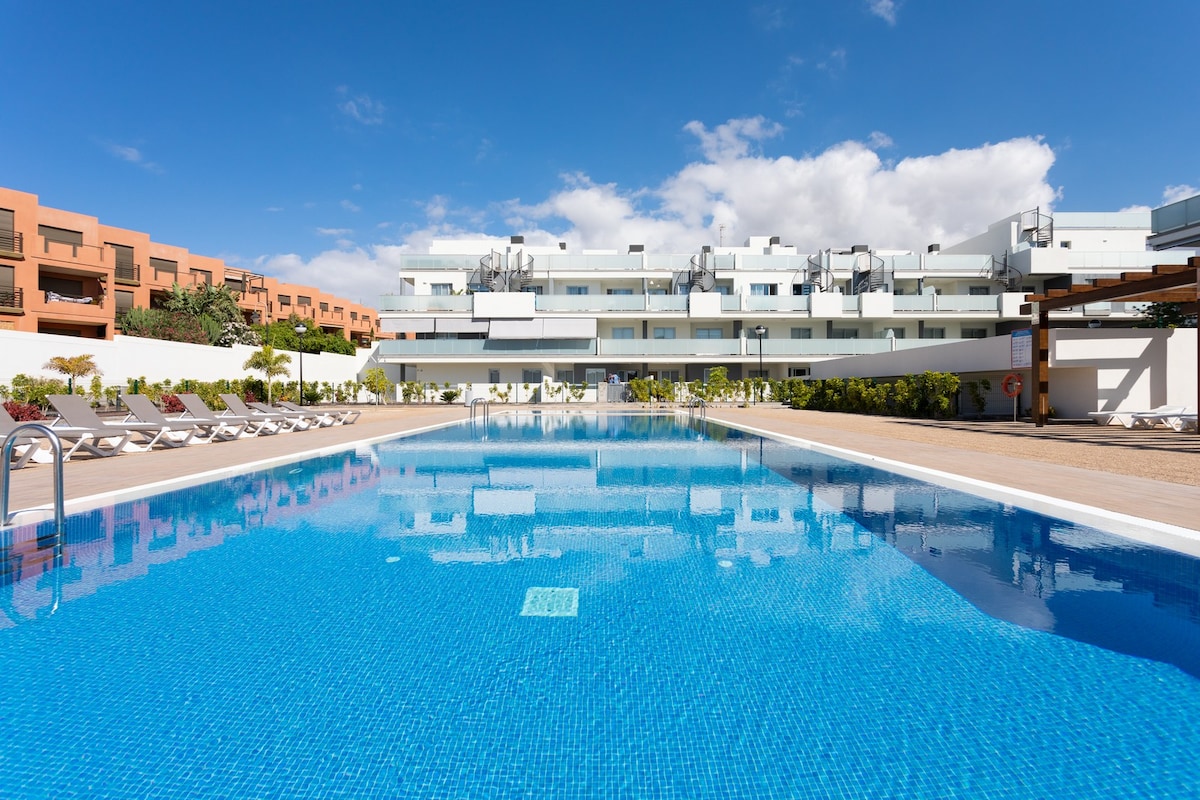 A clear blue swimming pool reflects the sky, with sun loungers positioned around its perimeter. The modern building can be seen in the background, featuring multiple balconies overlooking the pool area. Lush greenery adds a touch of nature to the surrounding space.