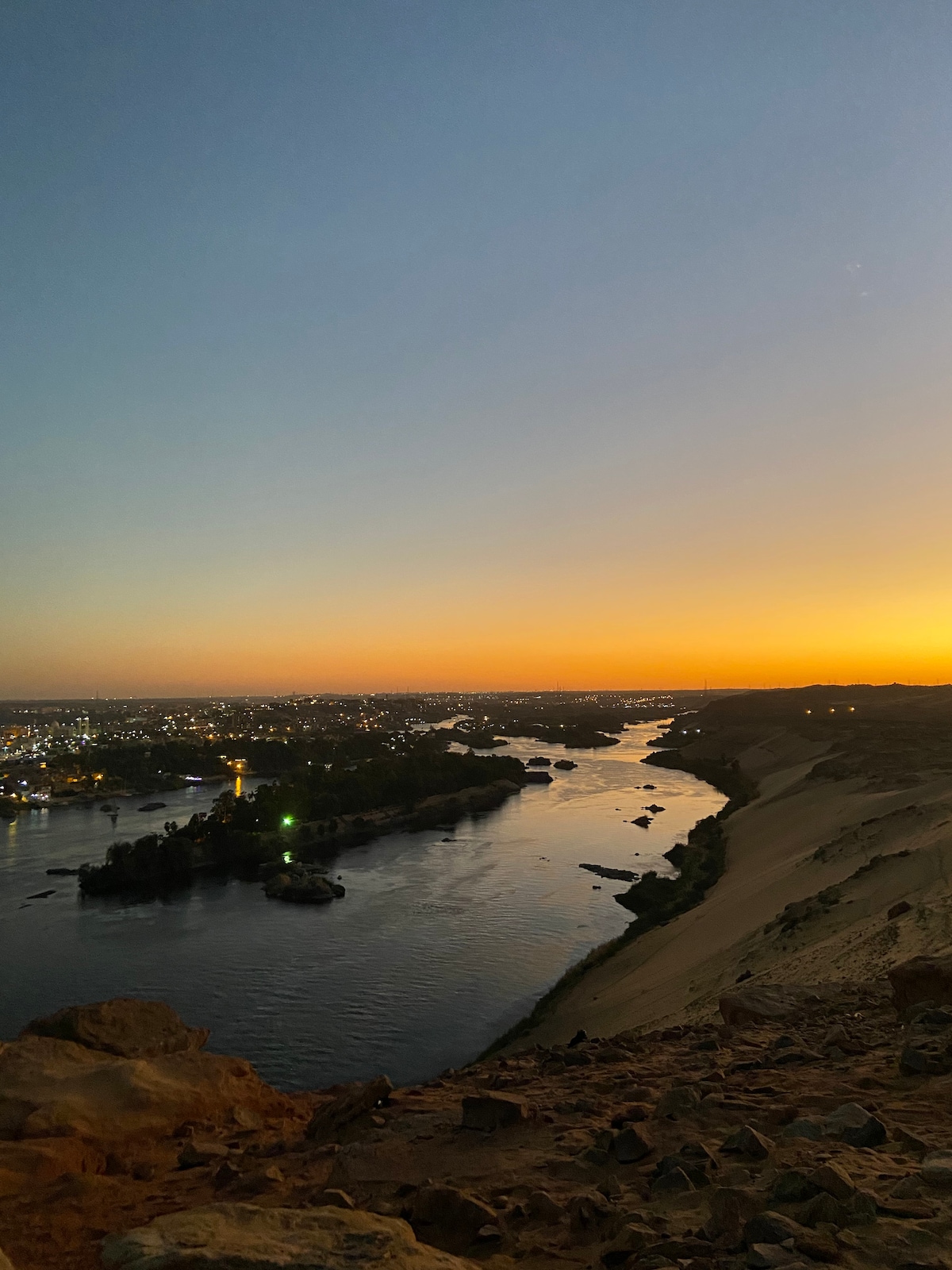 A serene view captures the Nile River flowing gently through the landscape, with islands dotting its surface. The horizon is painted in warm hues of orange and yellow as the sun sets, providing a tranquil backdrop to the village's gentle glow on the riverbank.
