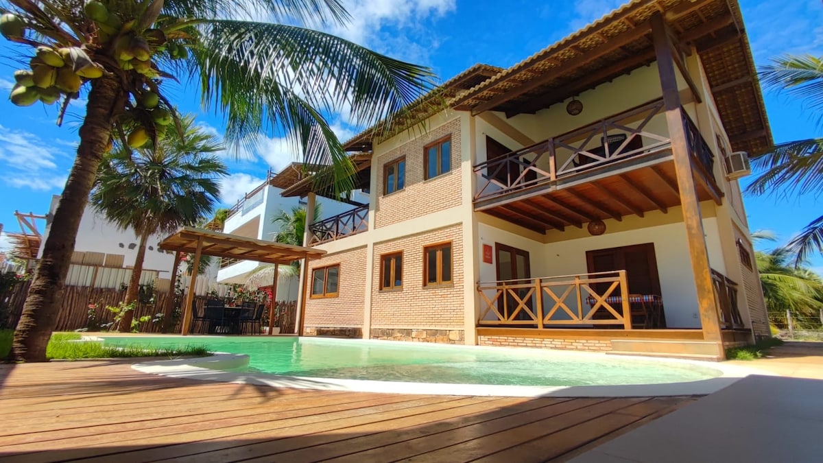 A three-story house is framed by palm trees, featuring a welcoming pool area surrounded by wooden decking. Expansive balconies with wooden railings are visible on the upper floors, while large windows allow for natural light to fill the interior spaces.