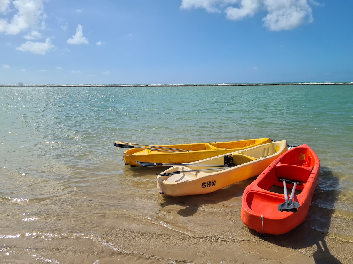Three kayaks are resting on the sandy shore, partially submerged in clear, turquoise water. The yellow kayaks are positioned gently beside a red one, with a backdrop of a blue sky scattered with light clouds.