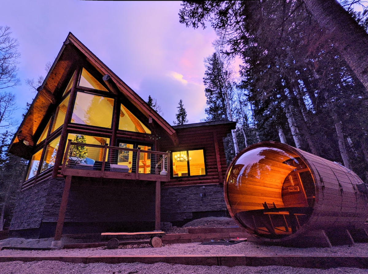 The cabin's exterior features a striking design with large windows reflecting warm light as dusk approaches. A barrel sauna sits nearby, crafted from wood and inviting, surrounded by tall trees and a tranquil forest setting.