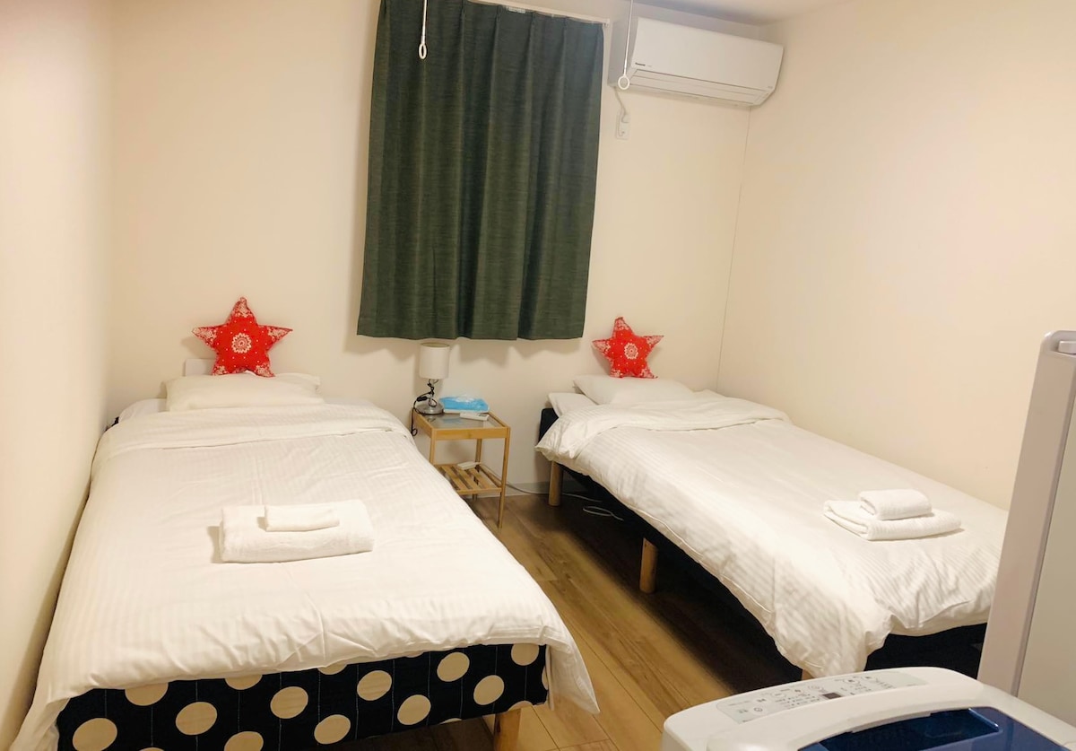 A bedroom containing two single beds, each adorned with red star-shaped decorative pillows. A small nightstand with a lamp is positioned between the beds. A window is draped with green curtains, and an air conditioning unit is mounted on the wall.