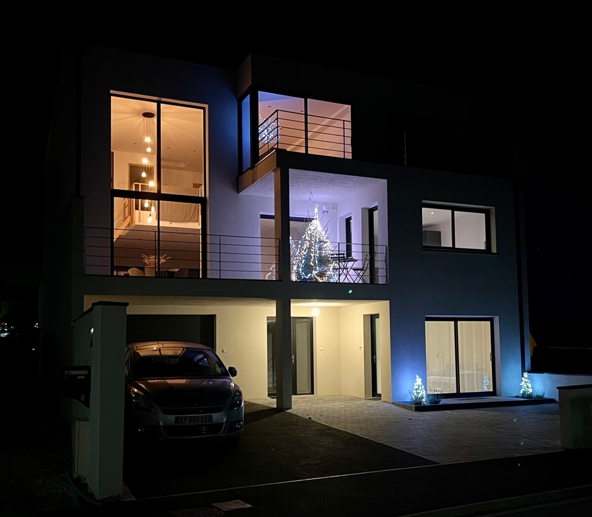 The modern house is illuminated at night, showcasing large windows that reveal a tastefully decorated interior. A Christmas tree is visible from the balcony, adding a festive touch. Soft lighting highlights the entrance, and two small trees are placed beside the front door.