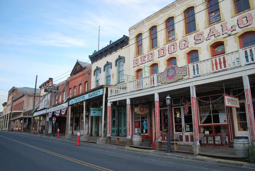 A historic street scene showcases a row of vintage buildings with various signage, including the 'Red Dog Saloon.' The architecture features distinct balconies and colorful façades, conveying a sense of heritage and charm. Flags and banners add a festive element to the storefronts.