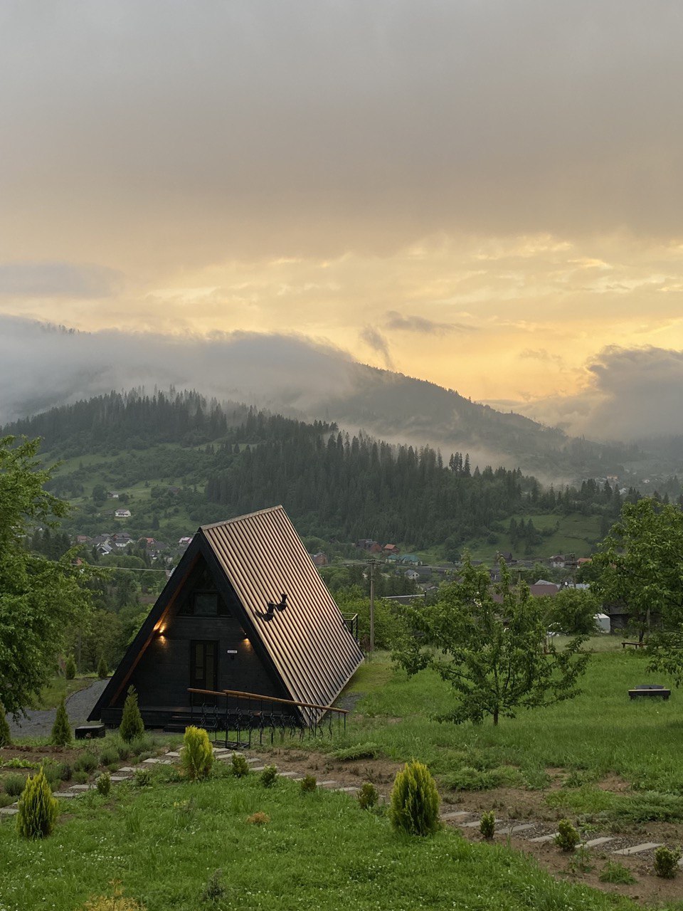 A modern cottage is positioned on a hillside, framed by lush greenery and rolling mountains. The sloped roof and black exterior contrast against the soft hues of a cloudy sunset, while the surrounding landscape blends seamlessly with the natural scenery.