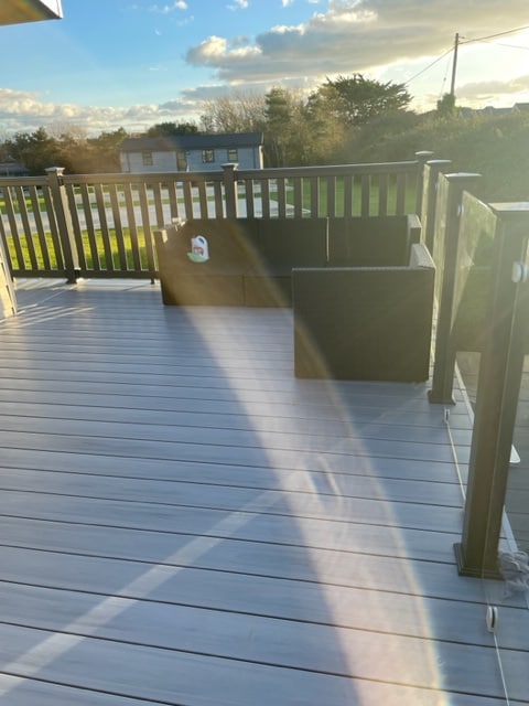 A spacious decking area features a rattan sofa set, surrounded by a railing that offers views of the greenery beyond. Soft sunlight casts gentle shadows, enhancing the surface's textures. The setting invites relaxation in a serene outdoor environment.