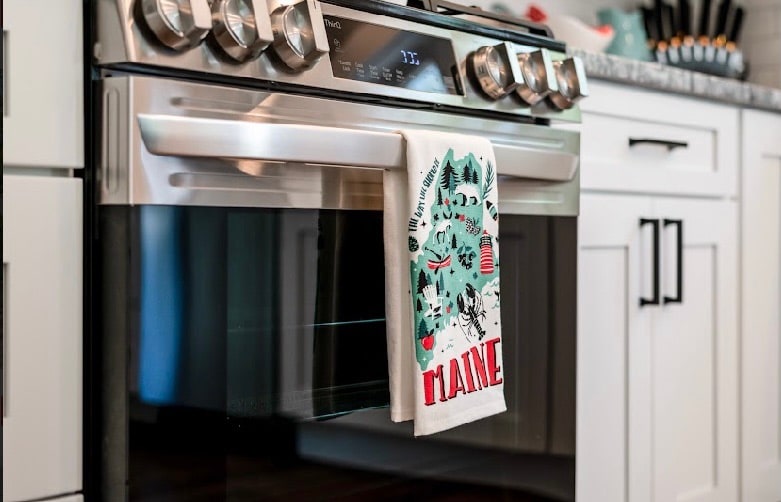 A modern stainless steel oven is positioned within a sleek kitchen. A decorative kitchen towel, featuring a design related to Maine, hangs from the oven handle. Light-colored cabinetry provides a clean and bright backdrop, with kitchen tools displayed in a holder on the counter.