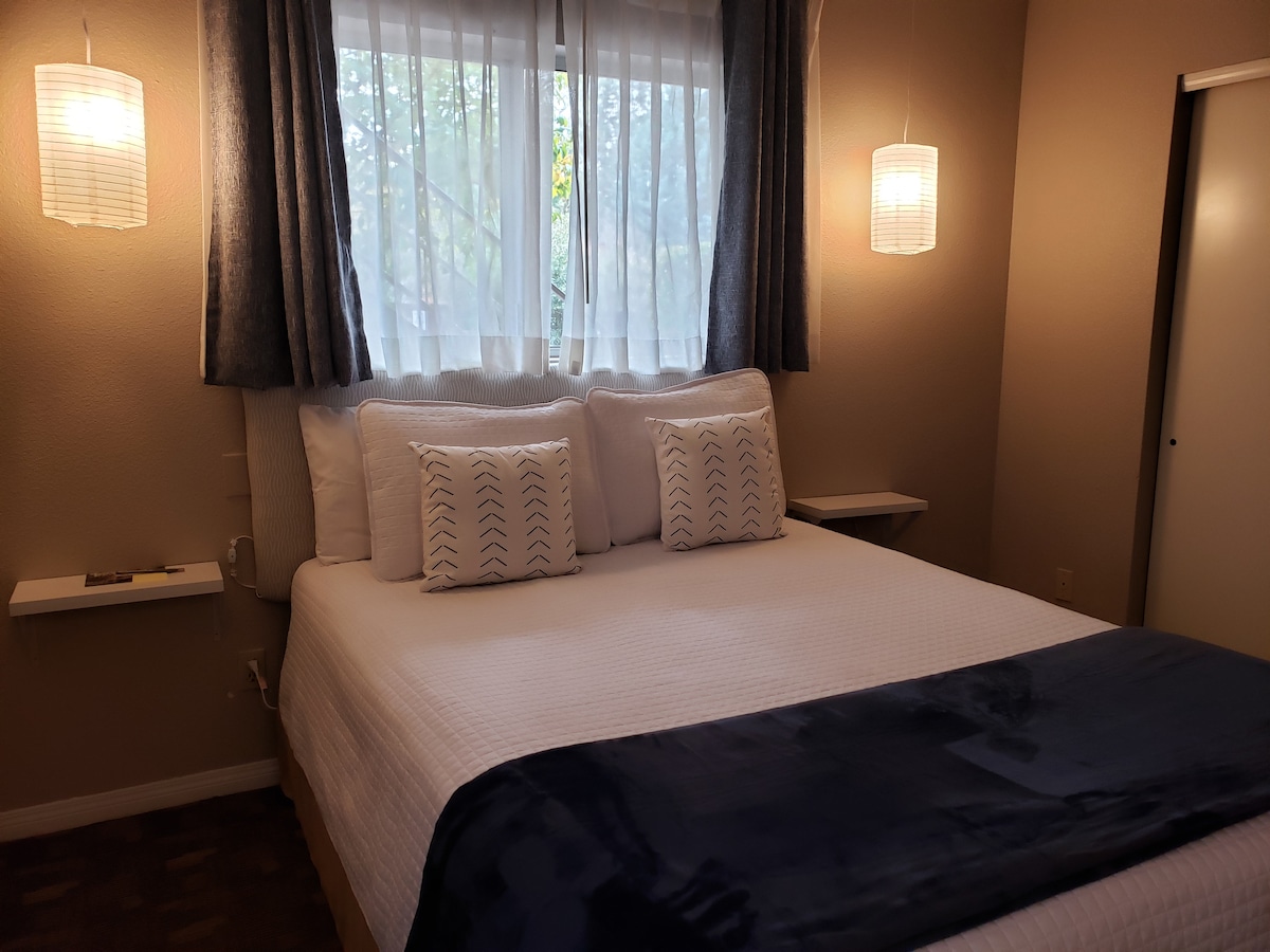 A cozy sleeping area is presented with a spacious bed, featuring white linens and decorative pillows. Soft lighting is provided by hanging fixtures on either side, while sheer curtains allow natural light to filter through the window. A small side table is positioned against the wall.