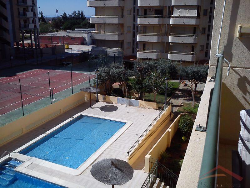 A panoramic view captures the outdoor pool, bordered by lounge areas and palm trees. Adjacent tennis courts are visible, alongside well-maintained green spaces. The surrounding residential buildings provide a serene backdrop to the leisure amenities offered in the community.