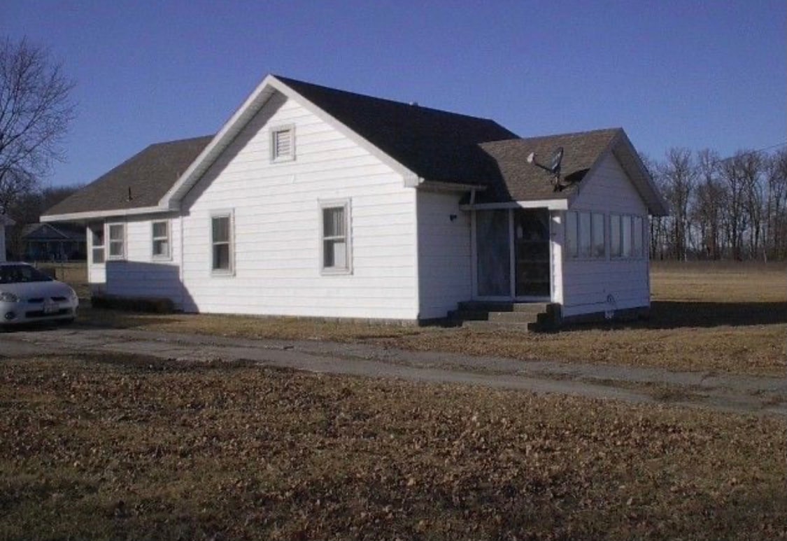 A single-story farmhouse is presented with a white exterior and a sloped roof. Large windows allow natural light to fill the rooms. The property is surrounded by open grass areas and trees in the background, providing a sense of space and tranquility.