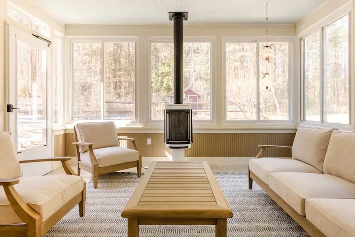 An enclosed three-season porch features large windows that provide natural light and views of the surrounding trees. Comfortable beige seating is arranged around a wooden coffee table, with a modern stove at the center, creating a cozy atmosphere for relaxation.