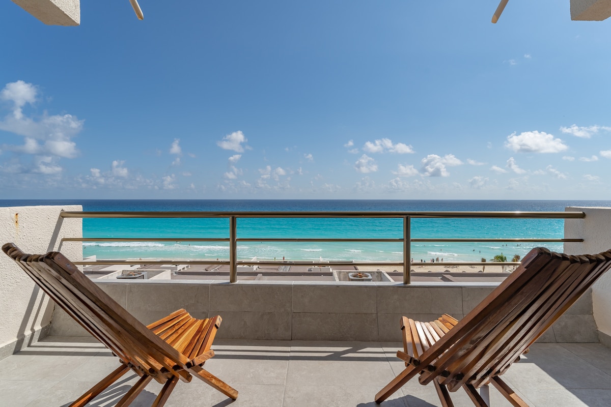 Two wooden lounge chairs are positioned on a balcony, providing a clear view of the Caribbean Sea. The turquoise water stretches towards the horizon under a bright blue sky with scattered clouds, creating a serene outdoor space for relaxation.