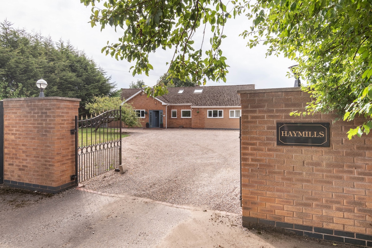 A gated entrance leads to a spacious gravel driveway surrounded by greenery. The brick house is partially visible, featuring multiple windows and a skylight. The sign 'HAYMILLS' is mounted on a brick pillar, indicating the property's name and welcoming guests.