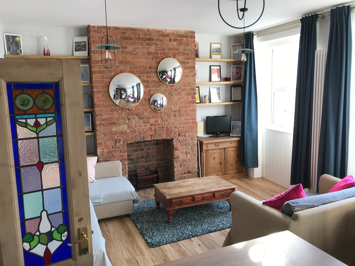 A living area features a brick accent wall adorned with three circular mirrors. A wooden coffee table sits on a textured rug, surrounded by a light-colored sofa and a blue armchair. Shelves with decorative items and a small television are positioned against the wall.