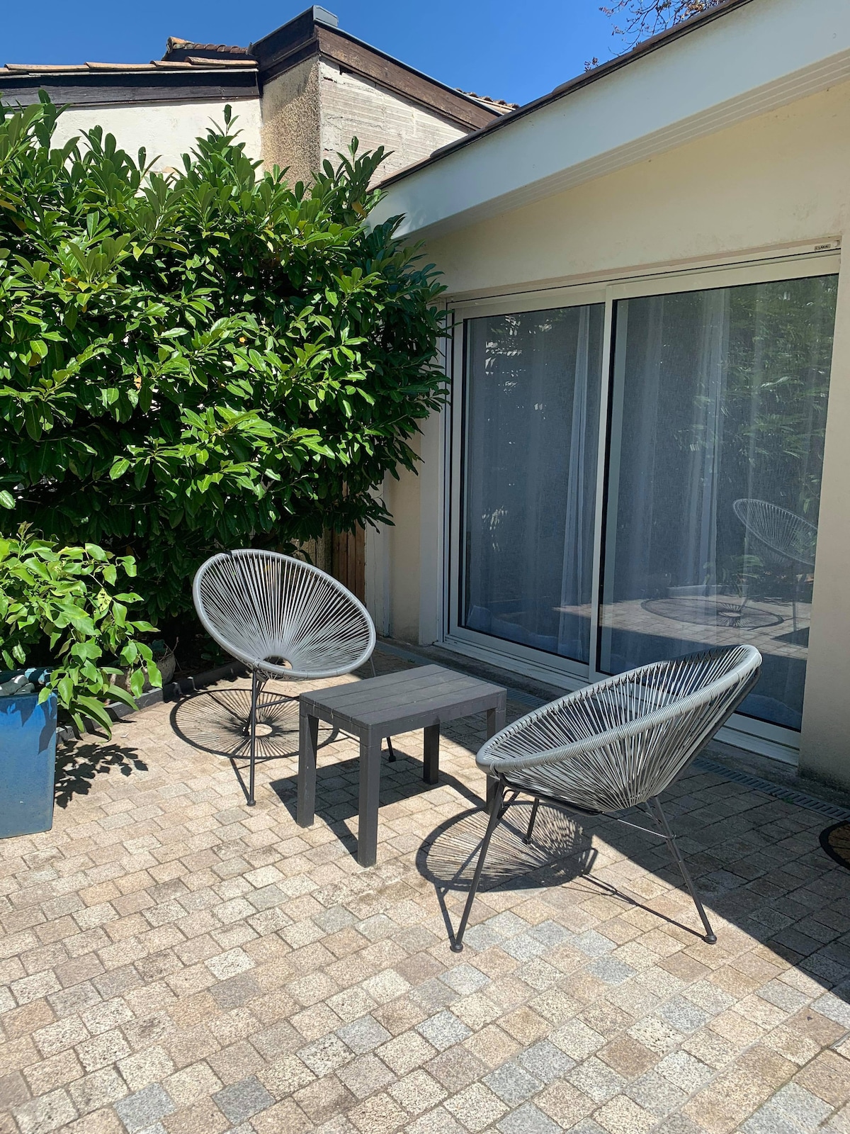 A private terrace features two woven chairs and a compact table, set on a stone-paved surface. Lush greenery provides a natural backdrop, while large sliding glass doors offer access to the interior space. Sunlight filters through, creating a bright and airy environment.
