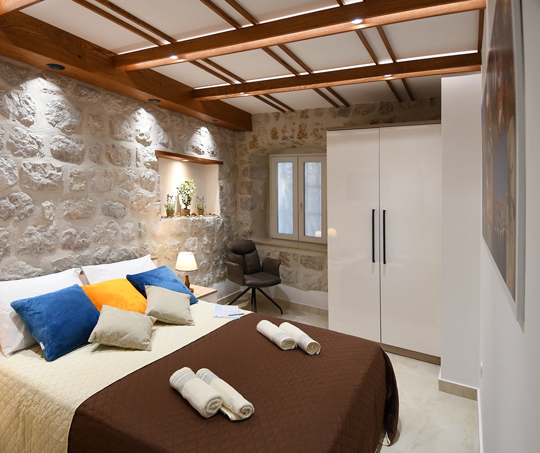 The bedroom features a double bed covered with a dark quilt and accented by colorful pillows. A modern white wardrobe stands against the stone wall. Soft lighting highlights the wooden beams above, while a minimalist chair sits near a window that overlooks the outside.