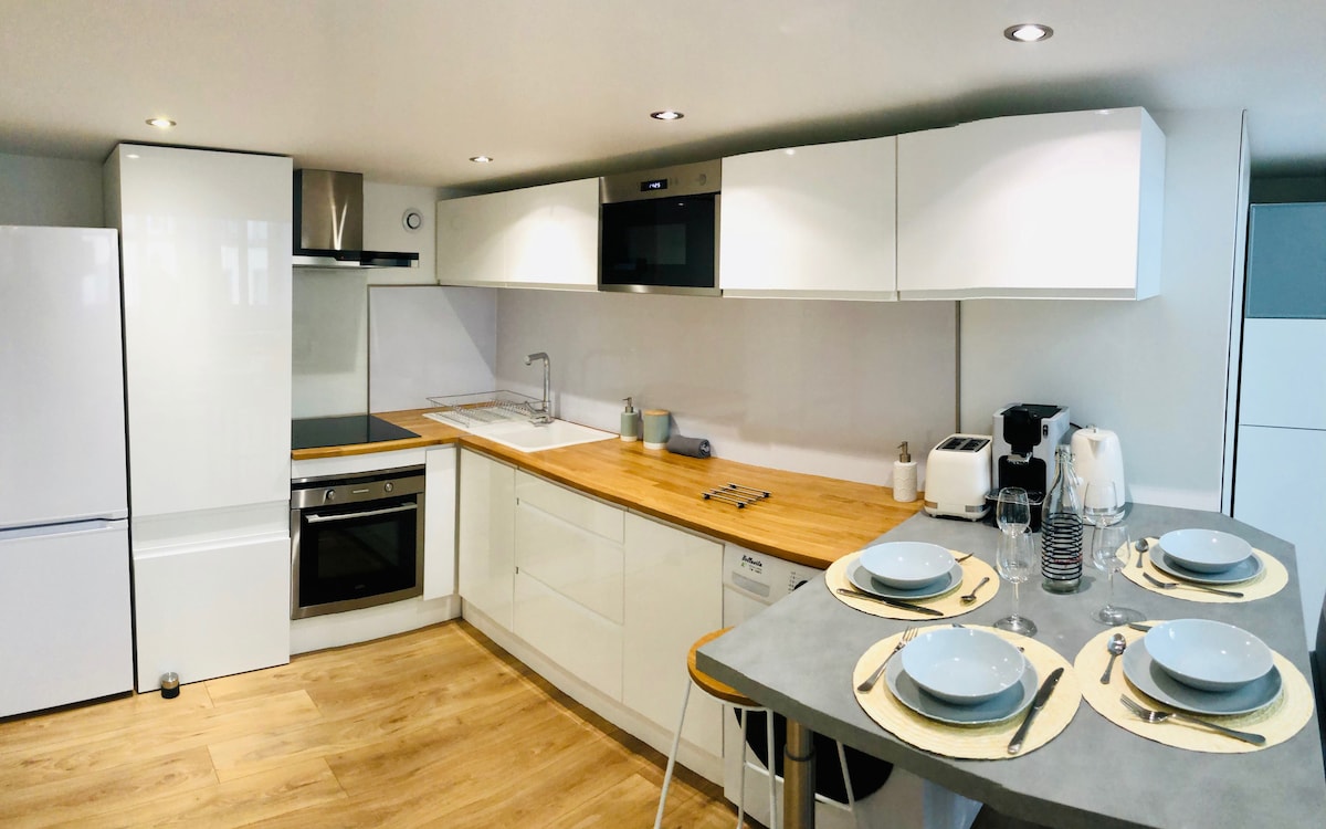 A modern kitchen featuring white cabinetry, a wooden countertop, and stainless steel appliances, including an oven and dishwasher. A dining table is set for four with plates, glasses, and cutlery. Warm wood flooring adds to the inviting feel of the space.