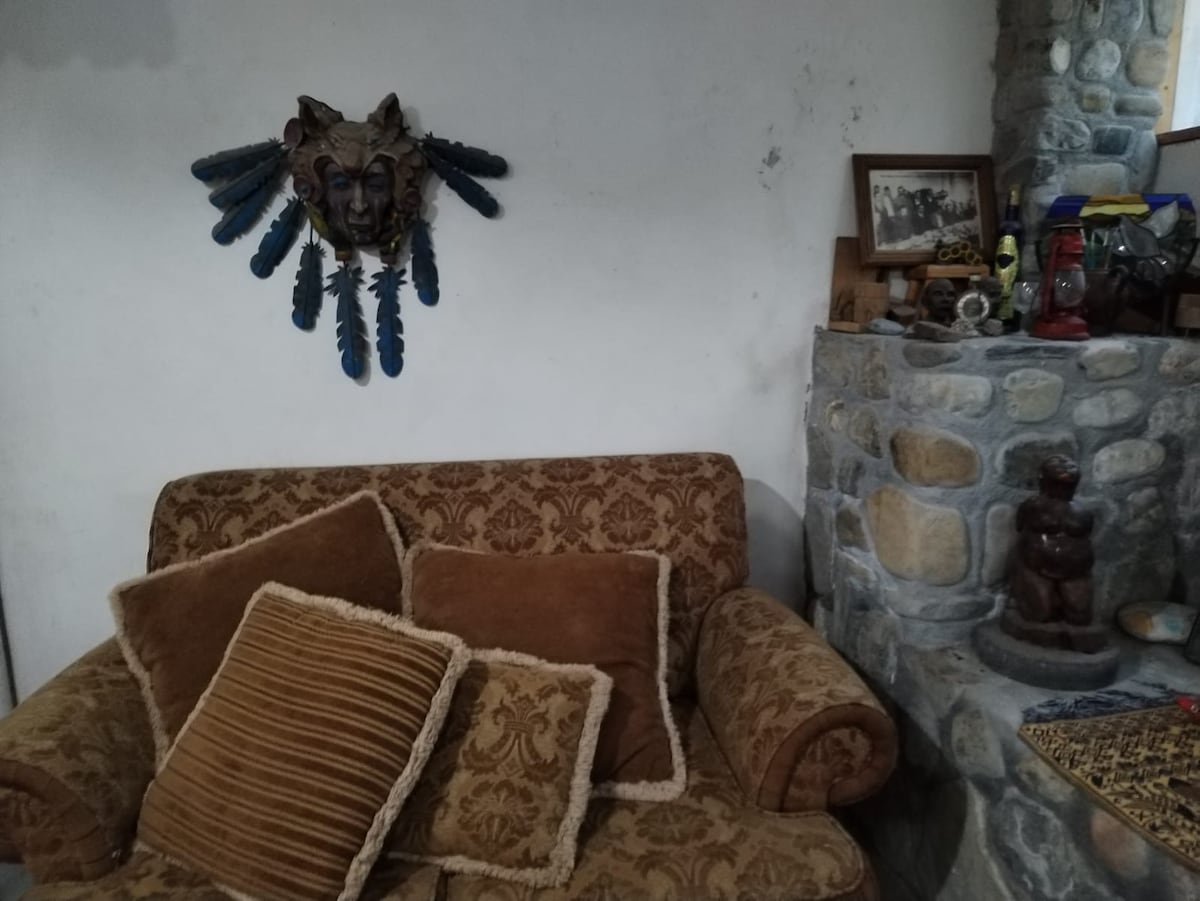 A patterned sofa is featured, accompanied by several cushions in varied textures. A decorative wall hanging with feathers is above the sofa, while a stone fireplace is positioned nearby, adorned with rustic decor and a framed picture.