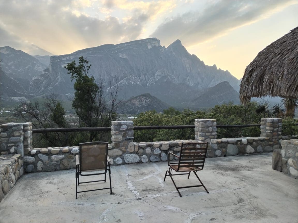 Two comfortable chairs are positioned on a stone terrace, offering unobstructed views of the surrounding mountains. The sky is adorned with soft, cloudy hues as the sun sets, creating a serene backdrop. A traditional palapa structure is visible to the side, enhancing the outdoor space.