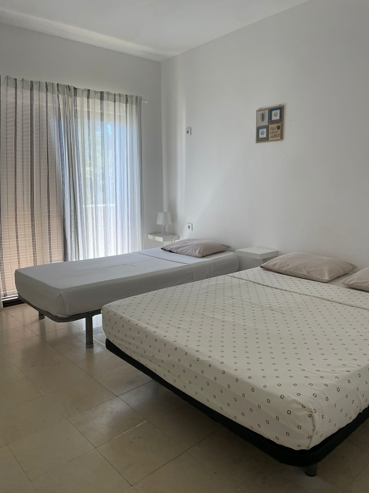 The room features a spacious layout with a double bed and a single bed, both equipped with simple bedding. Natural light filters through sheer curtains with a balcony door nearby. A few decorative frames are mounted on the white wall.