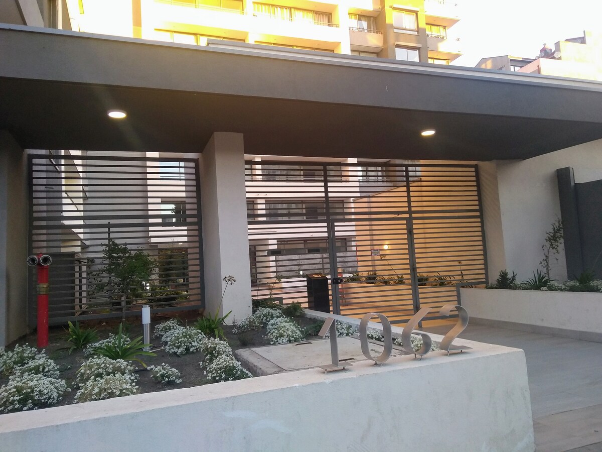 The entrance of a modern building is showcased, featuring a sleek awning and a landscaped area with white flowers. A large number '1062' is displayed prominently on the wall, and the surroundings include planter boxes and decorative greenery.