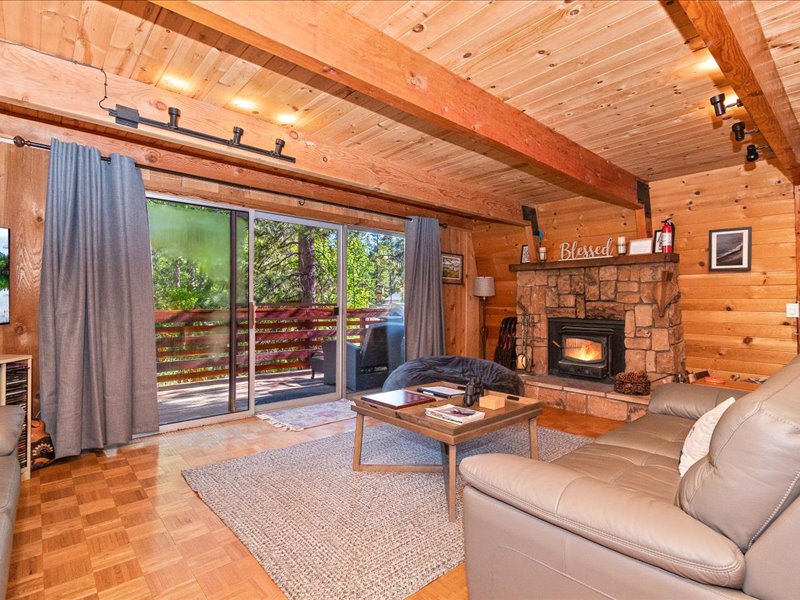 The living room features abundant natural light through large sliding glass doors, offering a scenic view of the outdoor deck. A stone fireplace enhances the space's warmth, complemented by wooden beams and cozy furnishings, including a soft rug and a coffee table.