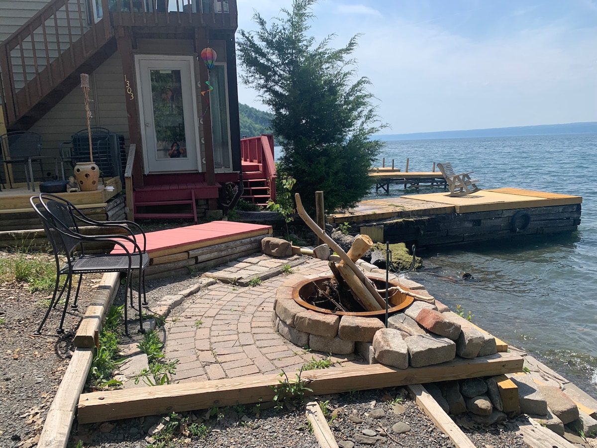 A stone fire pit surrounded by pavers is positioned near the water's edge, with two outdoor chairs placed invitingly beside it. A wooden dock extends into the lake, with views of the water and a tree providing some natural shade.