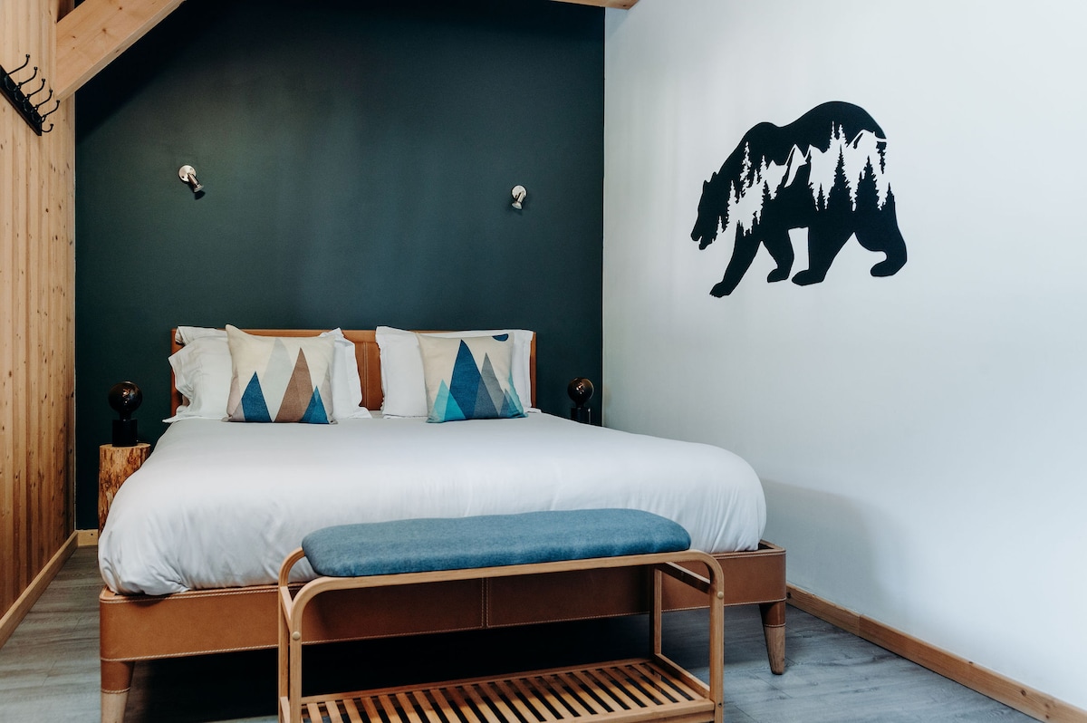 A spacious bedroom is characterized by a large bed with decorative pillows in geometric patterns. A wooden bench is placed at the foot of the bed. A mural of a bear silhouette adorns the wall, complemented by bedside lamps and warm wooden accents.