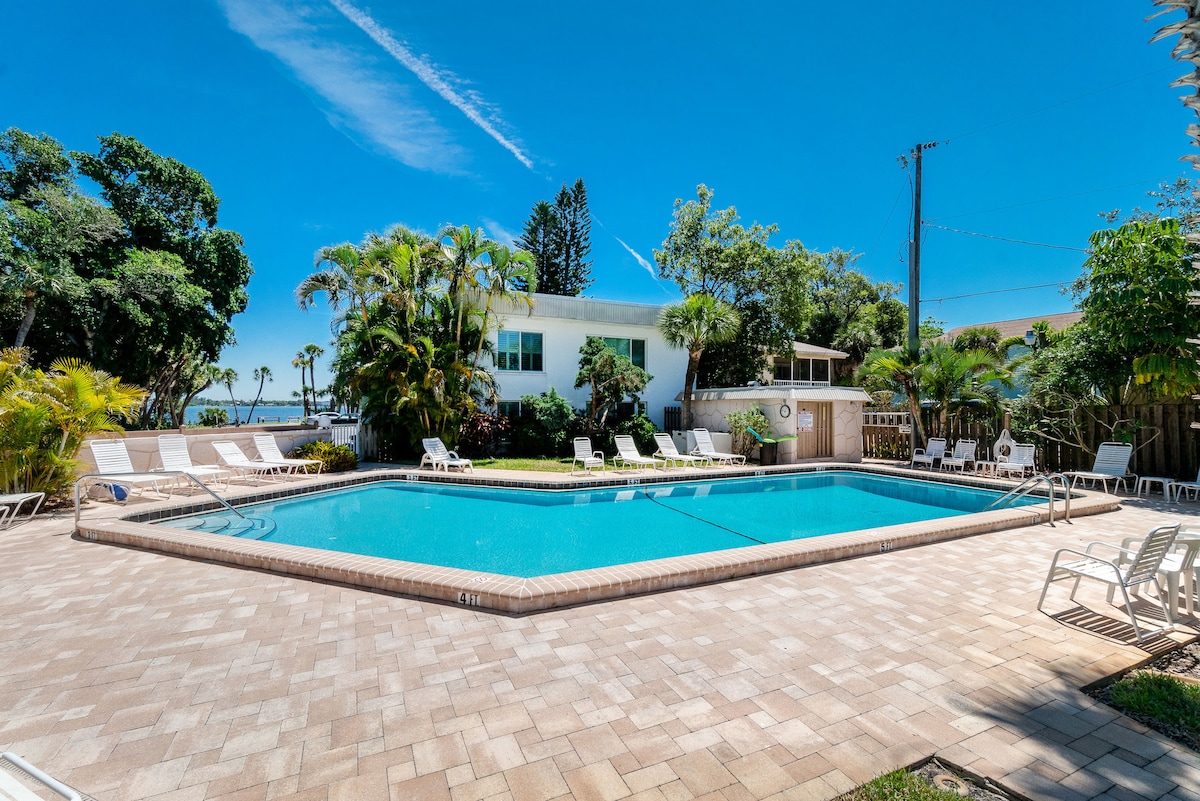 A spacious outdoor pool area is surrounded by tropical landscaping. Lounge chairs are arranged along the poolside, providing ample seating. The pool reflects a clear blue sky, with trees and a building visible in the background. The setting offers a relaxing environment for guests.