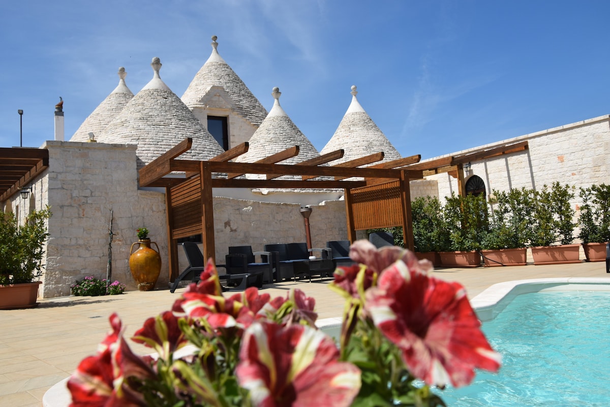 The exterior of a trulli structure is visible, featuring traditional conical roofs. A shaded seating area with dark furniture is positioned near a seasonal saltwater pool. Colorful flowers enhance the foreground, adding a vibrant touch to the natural stone surroundings.