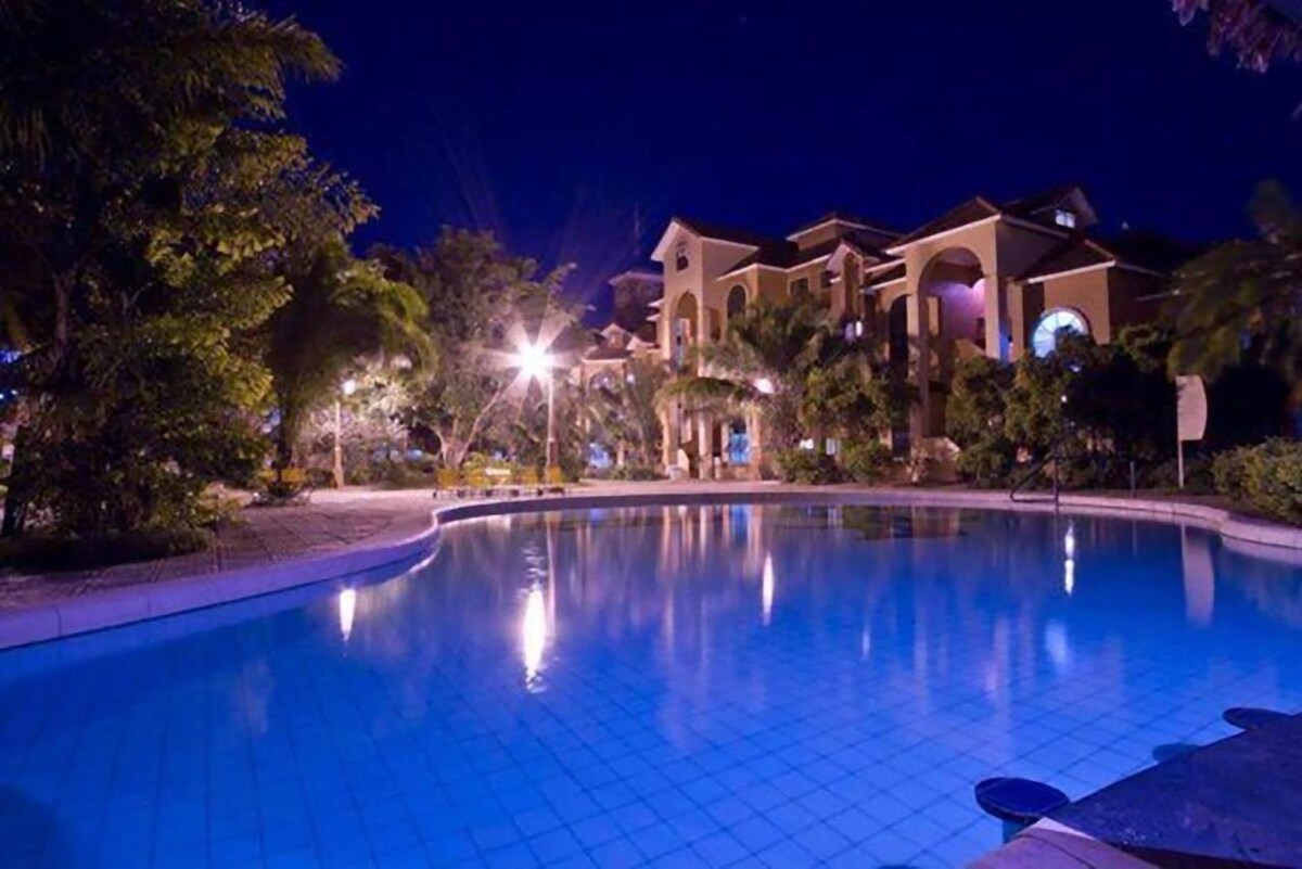 A well-lit swimming pool is surrounded by lush greenery, reflecting the soft glow of surrounding lights. The pool’s tranquil water invites relaxation, while an elegant building with multiple arches stands in the background, framed by palm trees under the night sky.