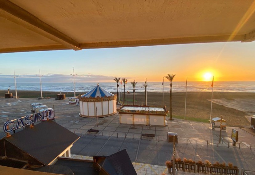 A sunset view overlooks the beach and ocean, with golden hues reflecting off the water. A carousel stands near the shoreline, framed by palm trees. The casino sign is visible in the foreground, with the tranquil atmosphere of the coast captured at dusk.