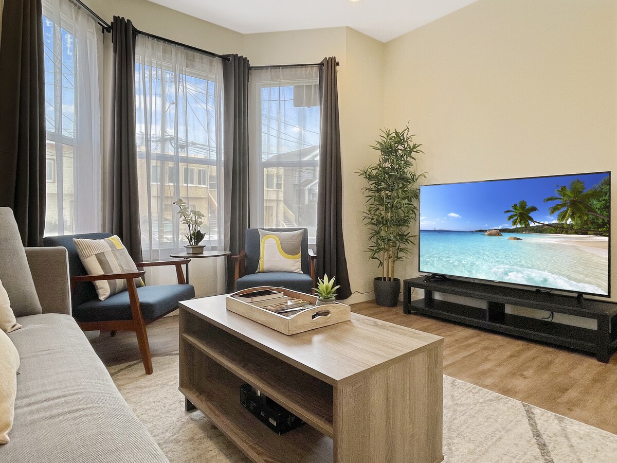 A bright living area features a cozy sofa and armchairs arranged around a wooden coffee table. Natural light streams through large windows dressed with curtains, while a TV displays a tranquil beach scene. A small plant adds a touch of greenery.