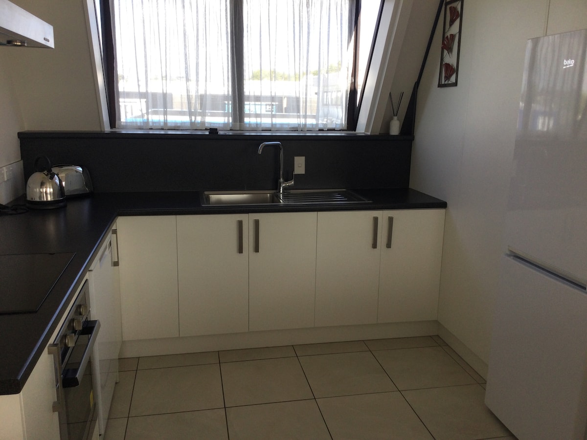 A modern kitchen presents sleek cabinetry and a compact layout. Natural light filters through large windows dressed in light curtains. A stainless steel kettle and a white refrigerator are visible, alongside an oven and a sink integrated into the counter space.