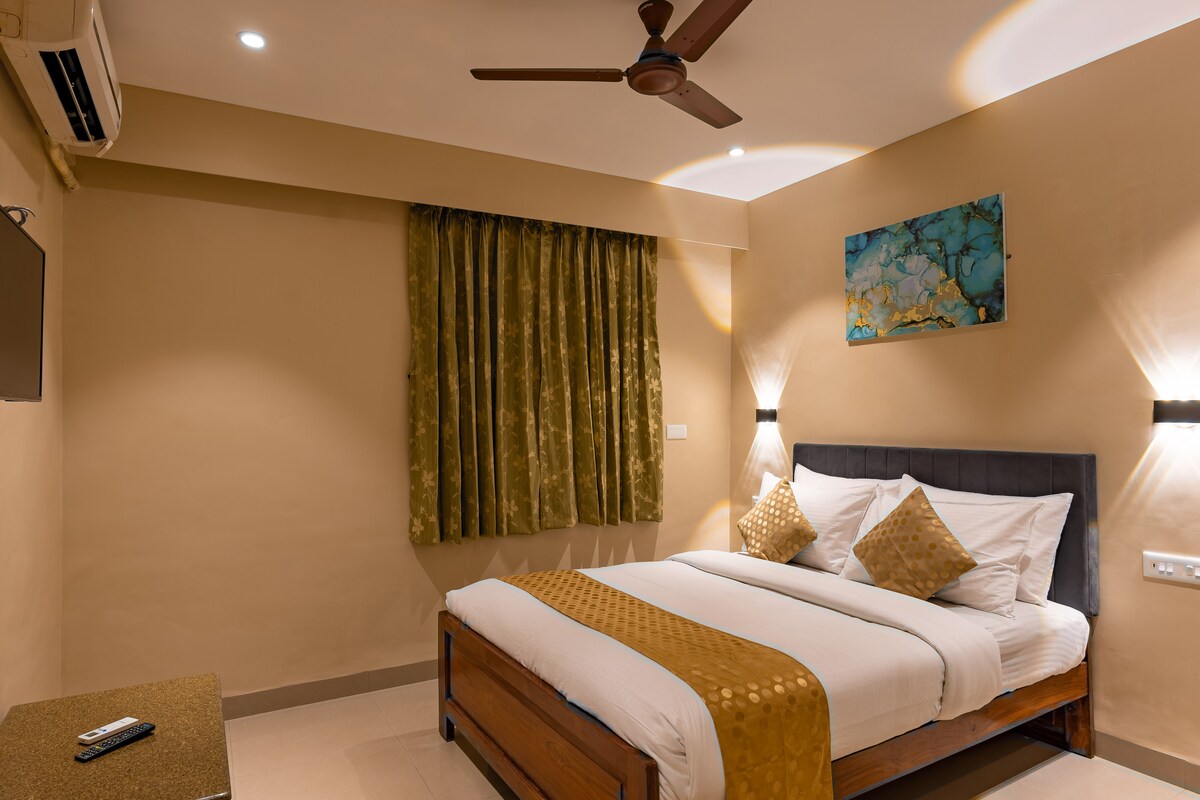 A cozy sleeping area features a spacious bed with decorative cushions, complemented by a soft bedspread. Two lamps provide gentle lighting, and a flat-screen TV is mounted on the wall. Dark curtains frame a window, adding a touch of elegance to the neutral-toned room.