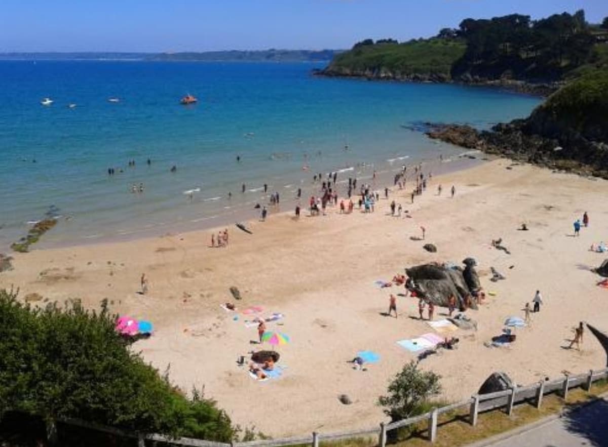 A sandy beach is presented, dotted with colorful umbrellas and beach towels. Groups of people are seen enjoying the sun and water, with gentle waves lapping at the shore. The backdrop features lush green cliffs under a clear blue sky.