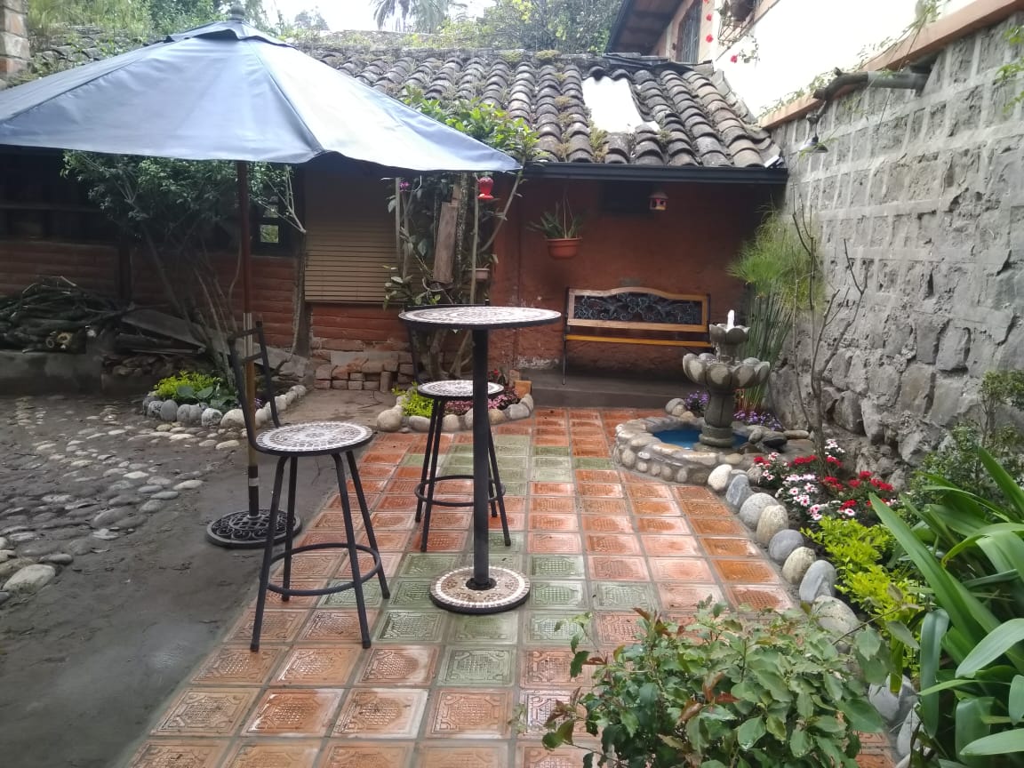 A charming garden area features a stone patio with decorative tiles, complemented by a small table and two chairs. An umbrella provides shade, while a stone fountain and colorful flower beds add to the serene ambiance of the space.