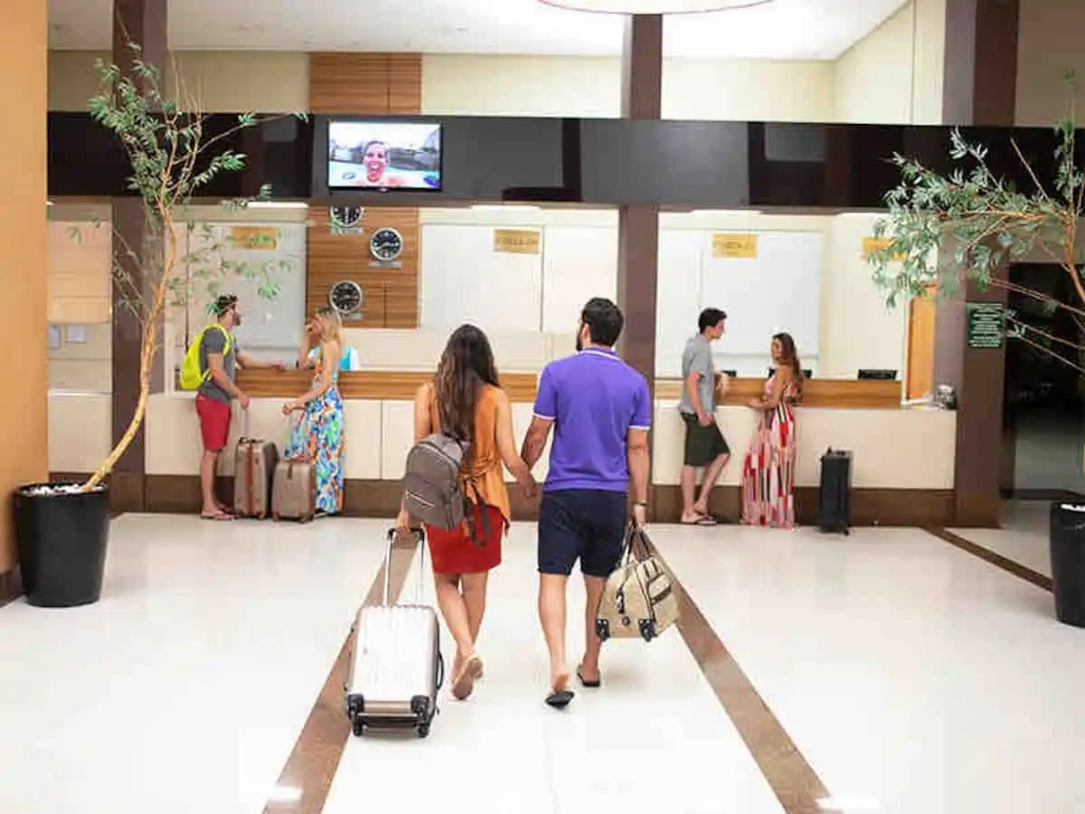 A spacious hotel lobby is featured, showcasing a reception area with several check-in counters. Guests with luggage are seen interacting with staff while others are gathered nearby. Decorative plants are placed around the space, and a television is mounted on the wall.
