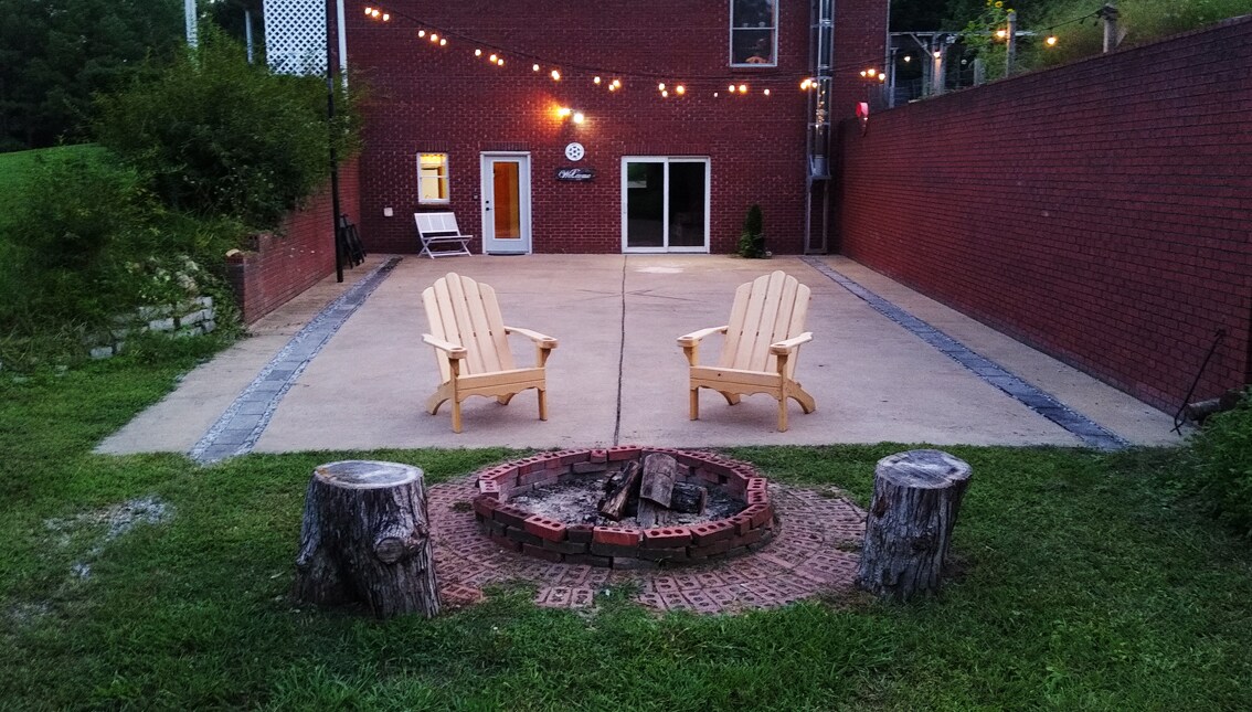 An outdoor patio area features two light-colored Adirondack chairs positioned around a circular brick fire pit, surrounded by timber stumps. Warm string lights are draped overhead, creating a cozy ambiance in this spacious setting, ideal for evening gatherings.