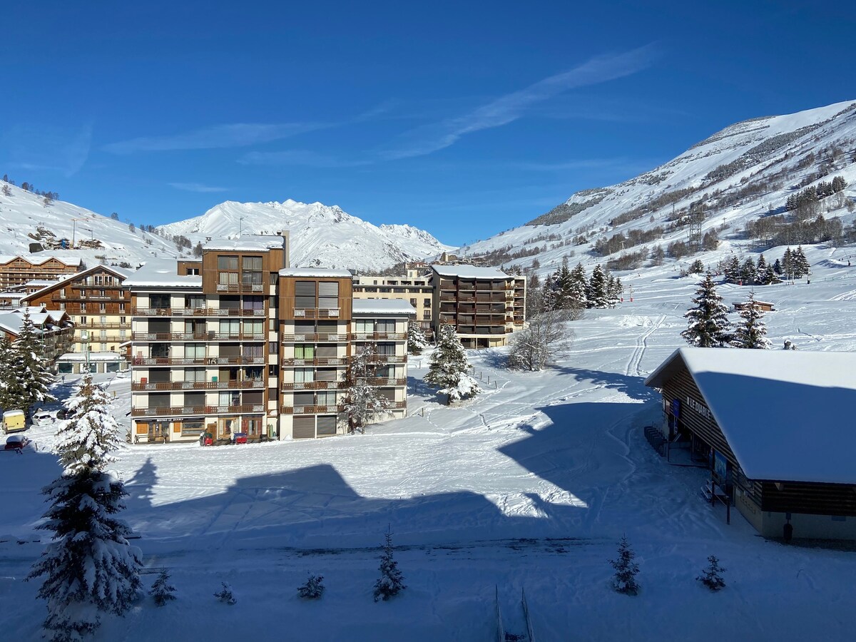 A snow-covered landscape is visible, featuring a modern multi-story residence amidst white slopes and pine trees. The bright blue sky provides a striking backdrop, while the nearby mountains create a scenic view reflecting the tranquil winter setting.