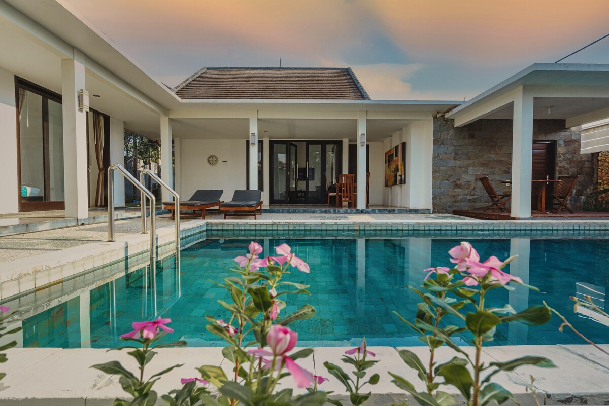 A private swimming pool is framed by blooming pink flowers, reflecting the serene atmosphere of the villa. The light-infused structure features a combination of sun loungers and a dining area, promoting an inviting space for relaxation and gathering.