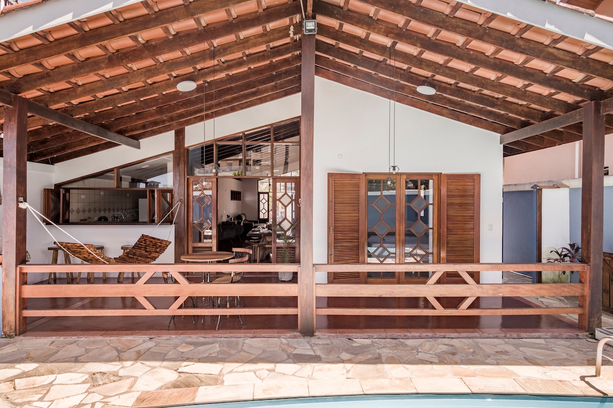 An open-plan living area features wooden beams and a high ceiling, enhancing the spacious ambiance. Large glass doors provide access to the outdoor space, while a hammock and dining area invite relaxation. Natural light brightens the interior, highlighting the stylish decor.