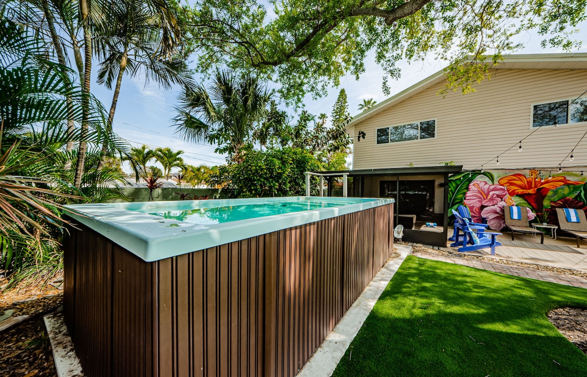 A large swim spa is surrounded by lush palm trees and tropical plants, creating a serene outdoor setting. The spa is positioned next to a spacious deck area, featuring colorful murals on the adjacent house. Green grass completes the inviting backyard scene.