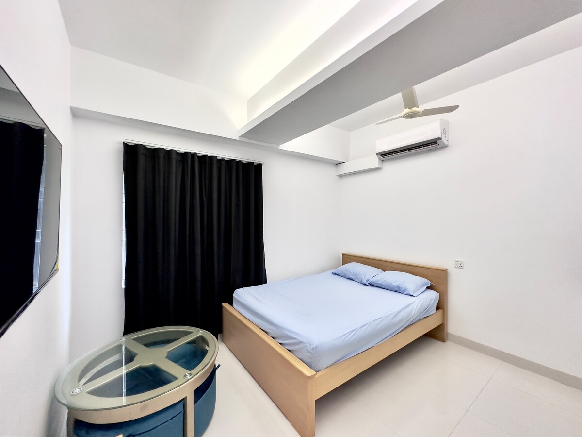 A bright studio apartment features a spacious bed with blue linens, complemented by a decorative glass table. A wall-mounted television is positioned opposite the bed, while a large window is covered by dark curtains, contributing to a restful ambiance.