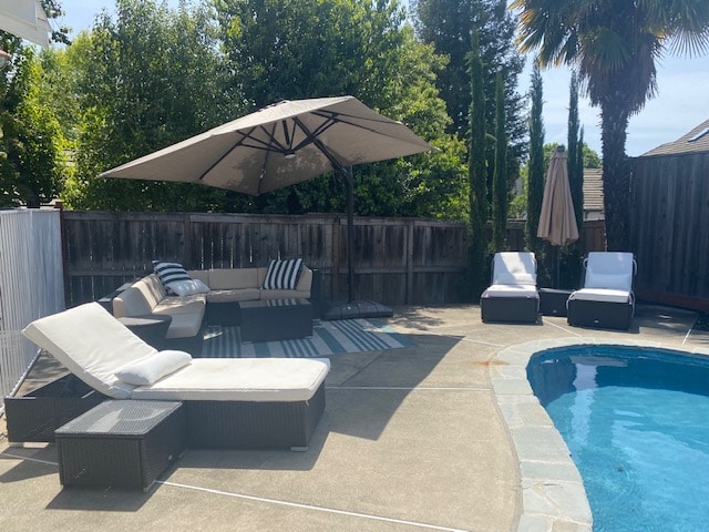 A spacious pool area features a solar-heated swimming pool surrounded by lounge chairs and an outdoor sectional. A large umbrella provides shade over the seating area, while a picnic table is visible nearby. Lush greenery surrounds the space, creating a relaxing atmosphere.